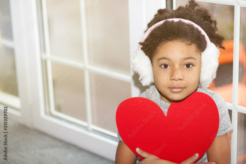 Adorable african american child girl with soft earphones holding a big red heart near the window. Love consept. Adorable african american child girl with soft earphones holding a big red heart near the window. Love consept.