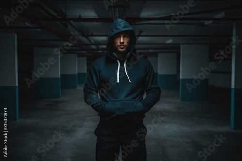 Serious attractive muscular caucasian sportsman in hoodie standing in underground garage with hands in pockets at night. Urban life concept.