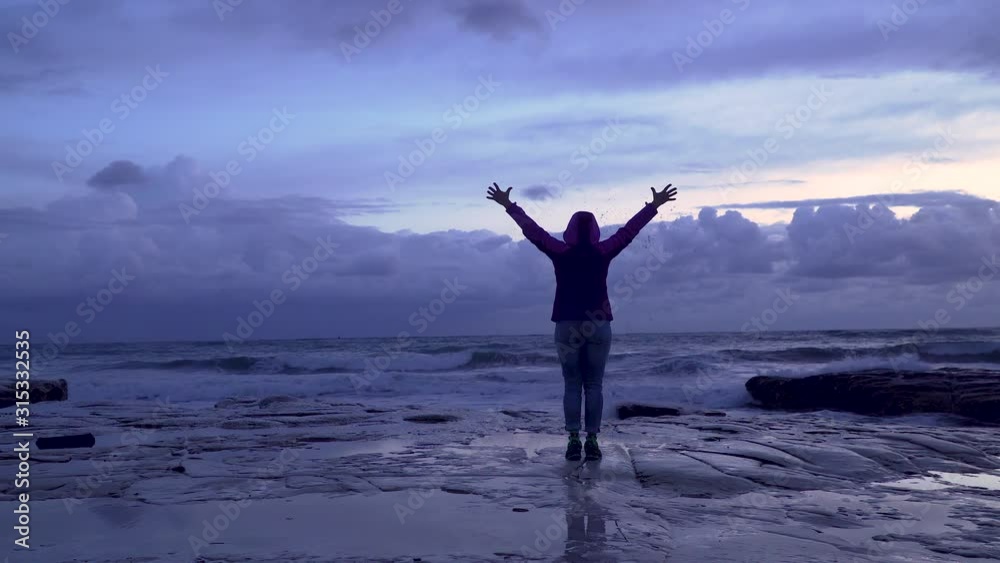 Girl on the seashore against the backdrop of storm waves. Woman raising arms victoriously. 4K Girl on the seashore against the backdrop of storm waves. Woman raising arms victoriously. 4K