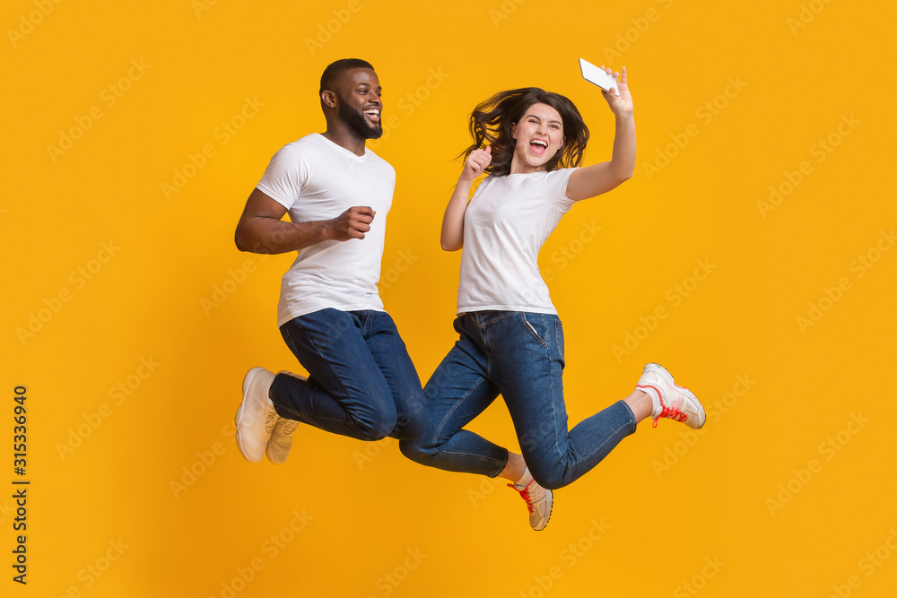 Joyful Interracial Couple Jumping In Air And Taking Selfie On Smartphone Joyful Interracial Couple Jumping In Air And Taking Selfie On Smartphone