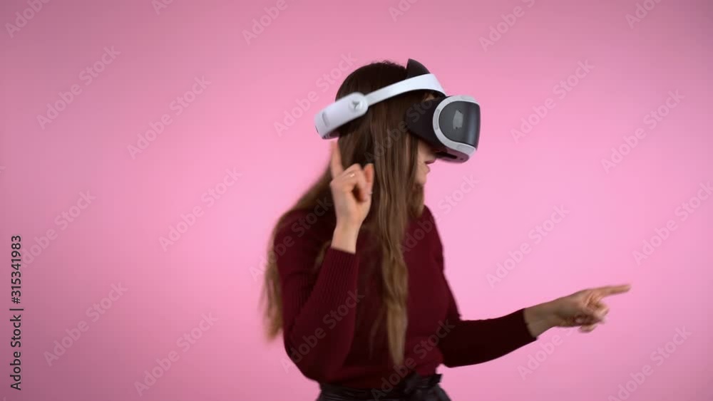 Moving gestures on pink background of happy young girl in virtual reality glasses headset. Engineering development, interactive innovations in electronics. Female playing games in augmented reality Moving gestures on pink background of happy young girl in virtual reality glasses headset. Engineering development, interactive innovations in electronics. Female playing games in augmented reality