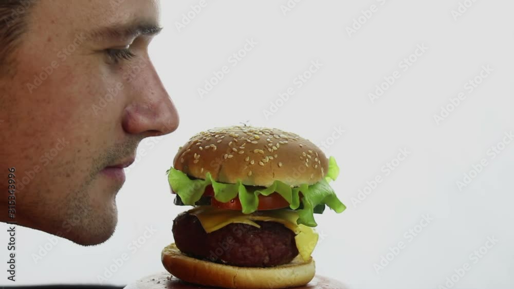 Young handsome guy sniffs and then bites a big juicy burger with great pleasure. Fast food, high-calorie food. Burger close-up on the white background. Young handsome guy sniffs and then bites a big juicy burger with great pleasure. Fast food, high-calorie food. Burger close-up on the white background.