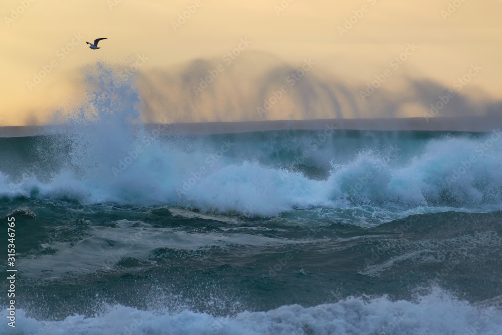 View of crushing ocean wave creating spiral spray trace on windy day in sunset light