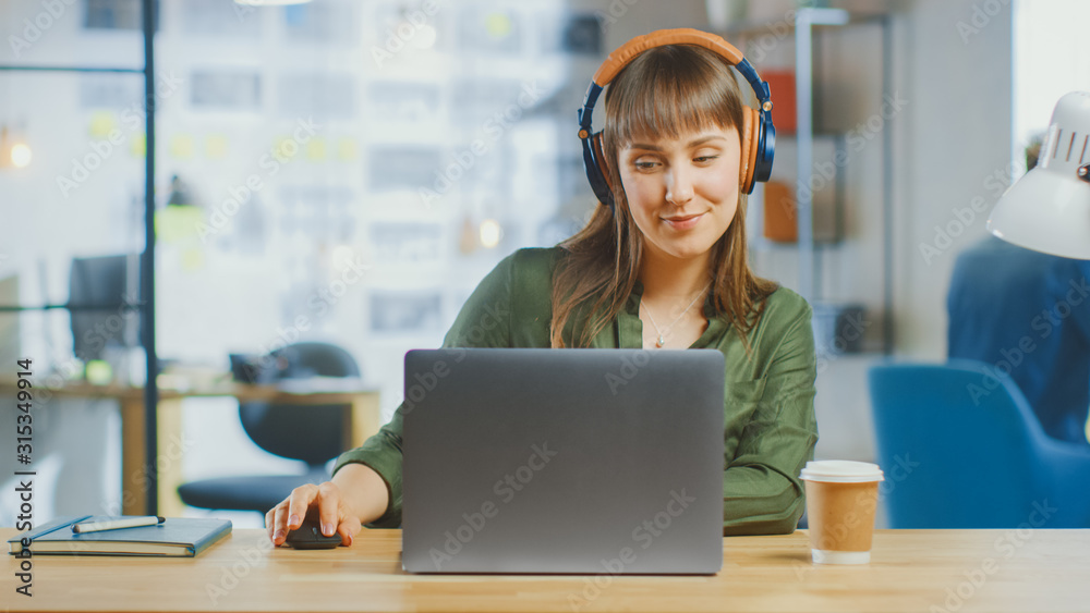 Young Beautiful Brunette Works on a Laptop Computer in Cool Creative Agency in a Loft Office. She is Wearing Headphones. She is Happy, Smiling and is Having Fun. Young Beautiful Brunette Works on a Laptop Computer in Cool Creative Agency in a Loft Office. She is Wearing Headphones. She is Happy, Smiling and is Having Fun.