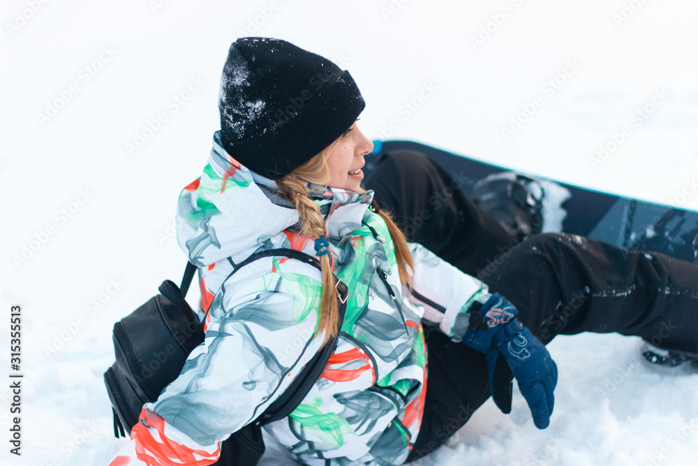 Beautiful blond girl in pigtails in colorful jacket with snowboard is sitting in profile on top of the mountain. Close up. Winter sunny day Beautiful blond girl in pigtails in colorful jacket with snowboard is sitting in profile on top of the mountain. Close up. Winter sunny day