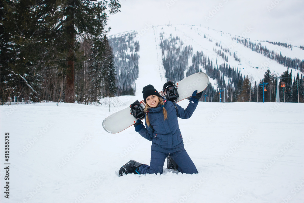 Beautiful young woman with the braids in grey winter costume is on her knees smiles and holds in the hands snowboard on a background of ski resort. Sunny winter day