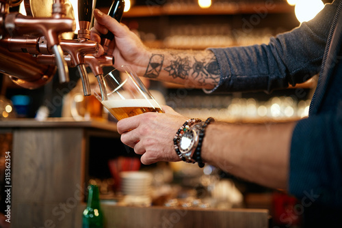 Tattooed caucasian barman pouring beer while standing in pub. Selective focus on hand.