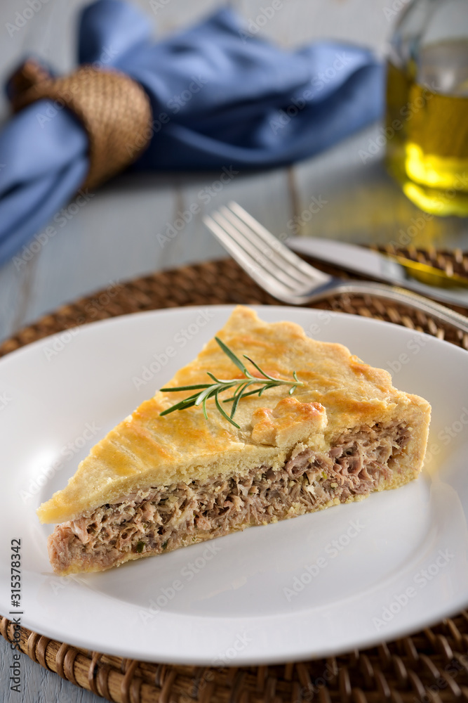 A slice of tuna fish pie with a crispy golden-brown crust, on a white plate with fork and knife on the background.