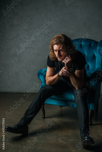 fashionable businessman in black outfit posing in armchair near grey wall