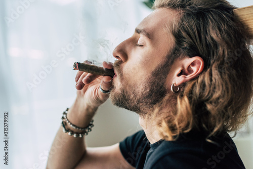 side view of fashionable casual businessman in total black outfit smoking cigar with closed eyes in office