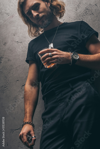 low angle view of fashionable businessman in black outfit with cigar and whiskey near grey wall