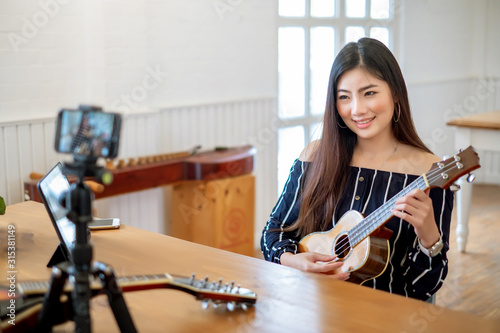 Asian woman blogger playing acoustic guitar recording video clip by smartphone and camera at home.