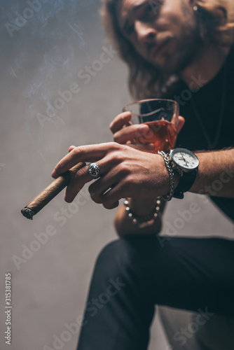 selective focus of fashionable businessman in black outfit holding cigar and glass of whiskey near grey wall