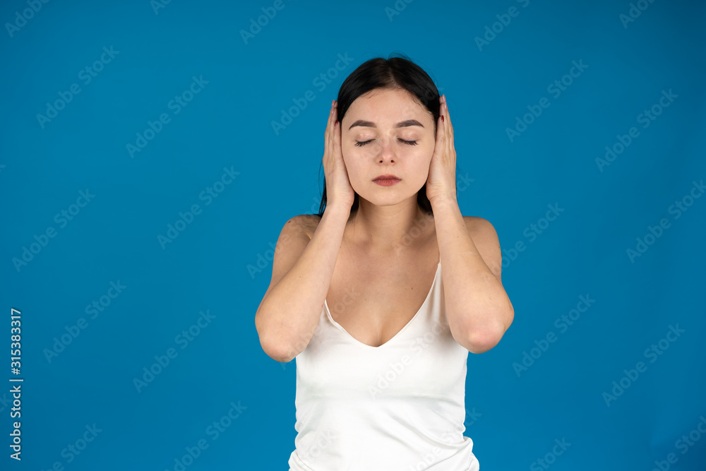 Front view of brunette girl with closed eyes covering ears with hands isolated on blue background Front view of brunette girl with closed eyes covering ears with hands isolated on blue background
