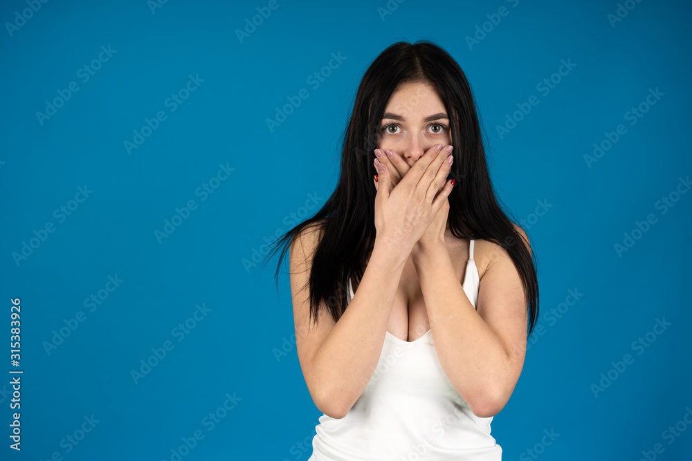 Front view of surprised woman covering mouth with hands isolated on blue background Front view of surprised woman covering mouth with hands isolated on blue background