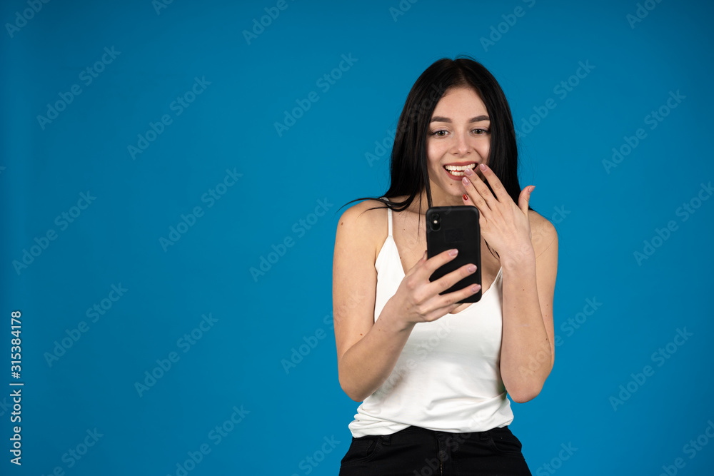 Front view of attractive girl covering mouth with hand while using smartphone isolated on blue background Front view of attractive girl covering mouth with hand while using smartphone isolated on blue background