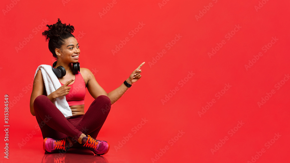 Cheerful fitness model sitting on floor, pointing at free space - obrazy, fototapety, plakaty 