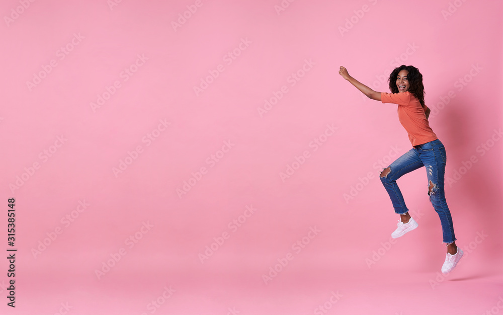 Joyful young african woman in orange shirt jumping and celebrating over pink banner background with copy space. Joyful young african woman in orange shirt jumping and celebrating over pink banner background with copy space.