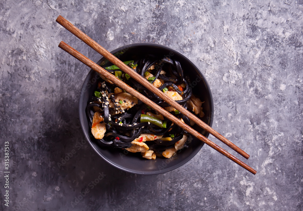 Black squid ink udon noodles with chicken meat in bowl with chopsticks on dark stone background. Top view. Black squid ink udon noodles with chicken meat in bowl with chopsticks on dark stone background. Top view.