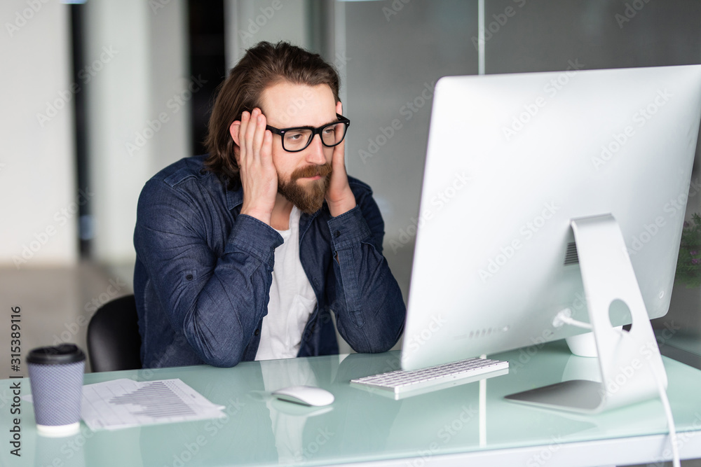 Tired or frustrated young man working in office looking at computer screen Tired or frustrated young man working in office looking at computer screen