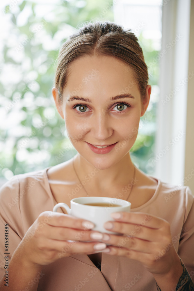 Vertical medium close up shot of charming young woman holding cup of tea looking at camera smiling
