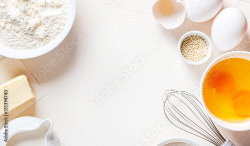 Frame of food ingredients for baking on a white background, top view, copy space. Flour, eggs, sugar and milk in white bowls and a whisk. Cooking and baking concept, flat lay.