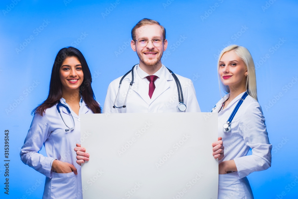 A team of young doctors with a sheet of white paper in his hands.. Multinational people - doctor, nurse and surgeon in blue background. A group of medical students of different nationalities A team of young doctors with a sheet of white paper in his hands.. Multinational people - doctor, nurse and surgeon in blue background. A group of medical students of different nationalities
