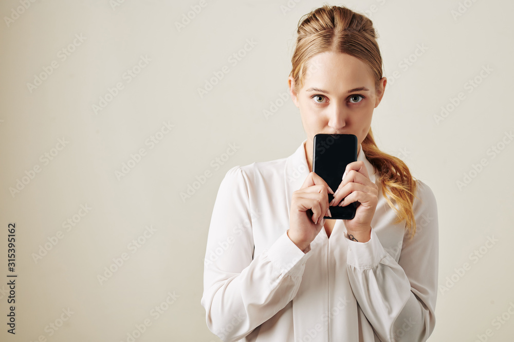Horizontal medium studio portrait of shy young woman wearing white blouse holding smartphoine with both hands Horizontal medium studio portrait of shy young woman wearing white blouse holding smartphoine with both hands