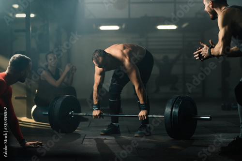 Male athlete exercising deadlift with barbell with support of his friends in a gym.