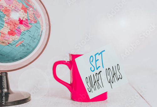 Conceptual hand writing showing Set Smart Goals. Concept meaning giving criteria to guide in the setting of objectives Globe map coffee cup sticky note lying vintage wooden table