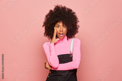 Amazed African American female wearing pink turtleneck and black overall touching face and looking at camera with open mouth surprised with news photo