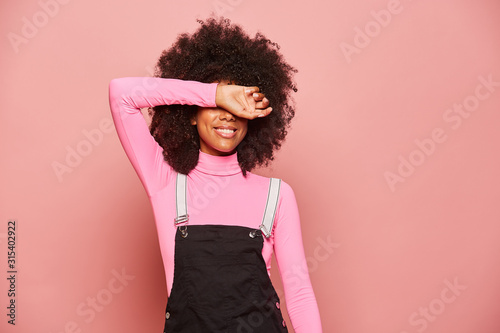 Excited millennial African American female with curly hair wearing pink turtleneck and black overall covering eyes with arm and waiting for surprise while standing against pink background photo