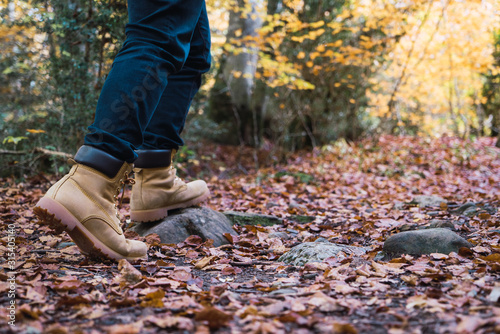 Crop legs in jeans and brown boots of hiker on rocky spangled of golden fallen leaves path with autumn forest on background photo