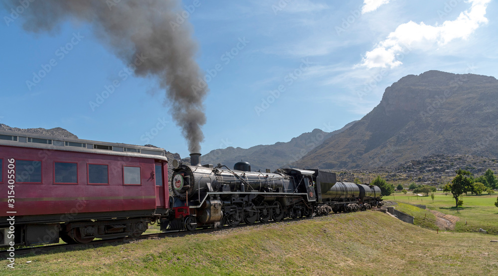 Obraz premium Ceres, Western Cape, South Africa. December 2019. Steam engine hauling passenger coaches to the annual excursion to the Cherry Festival on Ceres golf estate. Background of Michell's Pass.