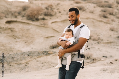 Cheerful bearded African American man in white t shirt and jeans holding newborn in baby carrier while strolling on nature