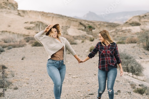 Woman in hat wearing casual clothes tenderly holding hands of female with long straight hair dressed in check shirt smiling on nature photo