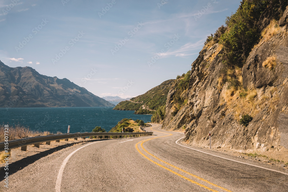 Asphalt road amid tropical green cliffs and blue lake with cloudy blue sky and mountain Cook on the background Asphalt road amid tropical green cliffs and blue lake with cloudy blue sky and mountain Cook on the background