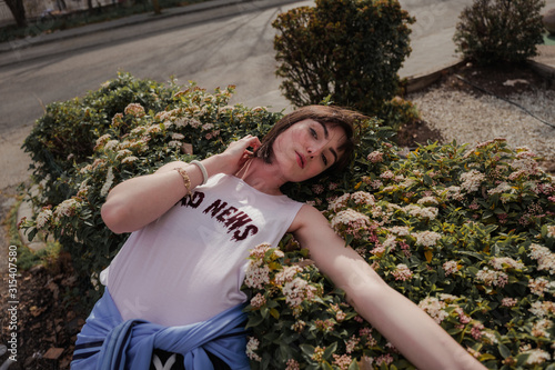 From above of elegant female teenager in casual stylish wear leaning on bloomy green shrub and looking at camera at city street photo