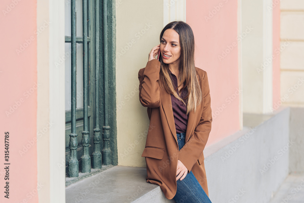 Satisfied charming lady in stylish casual clothing and heels leaning on wall of building talking on the mobile phone looking away at city street