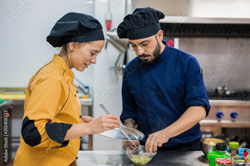 Young female cook in yellow uniform holding plate and tongs and male colleague in dark blue uniform holding bowl with food while working together in kitchen and preparing delicious dish