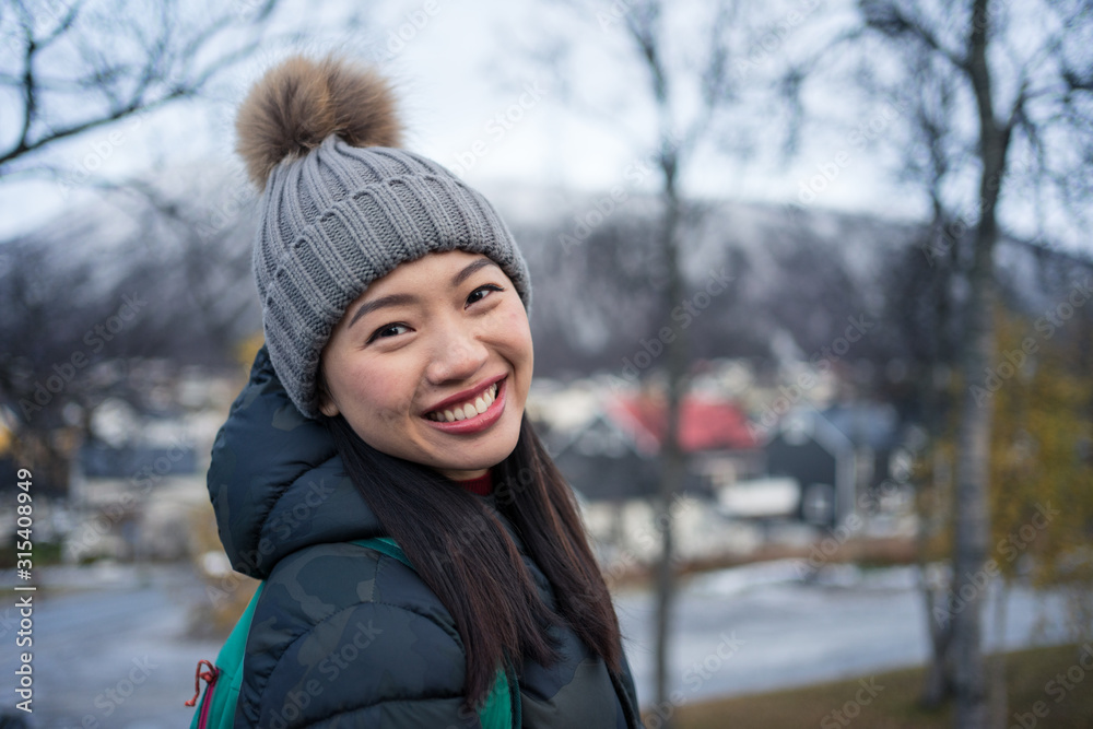 Attractive adult Asian woman in warm clothes with backpack smiling at camera while standing on street against blurred exterior of amazing triangle shaped church and snowy hills in Norway Attractive adult Asian woman in warm clothes with backpack smiling at camera while standing on street against blurred exterior of amazing triangle shaped church and snowy hills in Norway
