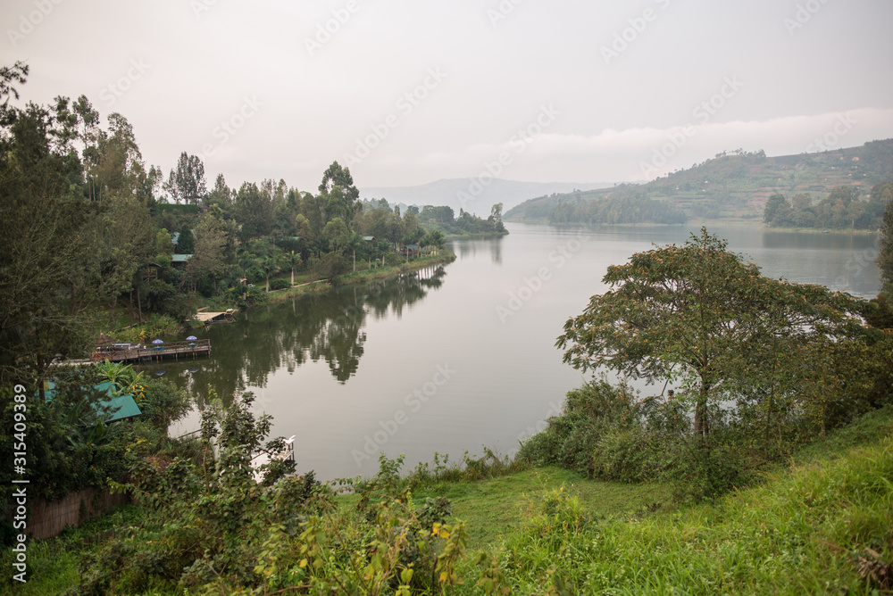 Uganda - November 26, 2016: Picturesque wild shoreline of wide quiet river with tranquil clear water among lush colorful bushes and green deciduous trees against foggy forested hills at horizon under cloudy sky in daytime Uganda - November 26, 2016: Picturesque wild shoreline of wide quiet river with tranquil clear water among lush colorful bushes and green deciduous trees against foggy forested hills at horizon under cloudy sky in daytime