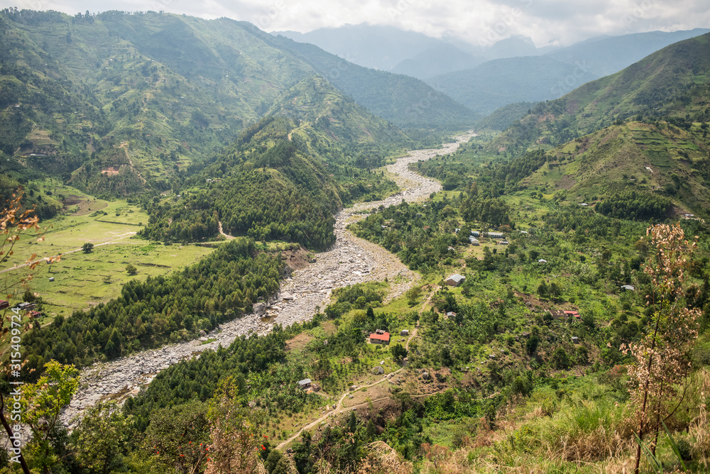 Uganda - November 26, 2016: From above magnificent scenery of countryside with small cultivated fields and solitary village along winding river in misty valley among hills with green lush grass and dense forests Uganda - November 26, 2016: From above magnificent scenery of countryside with small cultivated fields and solitary village along winding river in misty valley among hills with green lush grass and dense forests