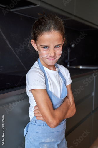 Little girl with blue eyes in apron with flour on cheeks standing with crossed hands and looking at camera pissed in a modern kitchen