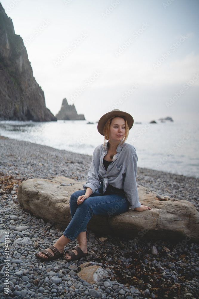 Dreaming stylish female tourist enjoying seascape while chilling on big stone on rocky shore of Asturias looking away Dreaming stylish female tourist enjoying seascape while chilling on big stone on rocky shore of Asturias looking away