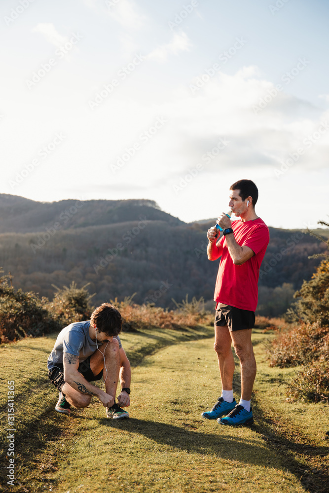 Adult athletic man standing on green trail and drinking water while friend tying running shoes during training in mountains in sunny day Adult athletic man standing on green trail and drinking water while friend tying running shoes during training in mountains in sunny day