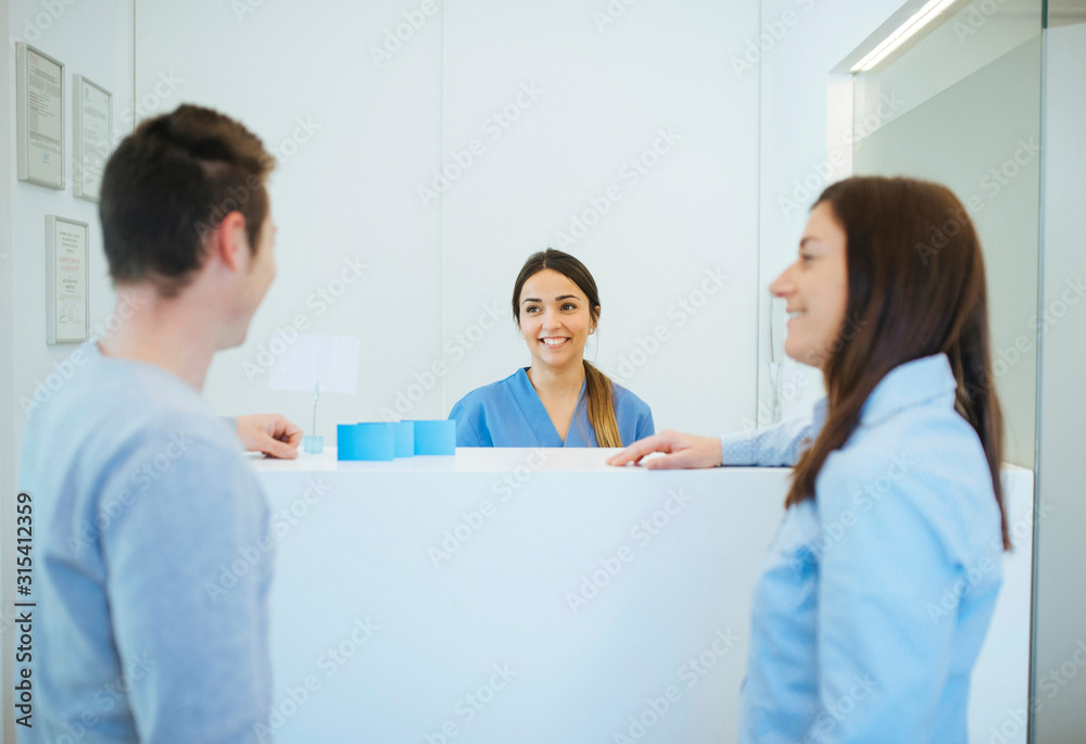 Side view of smiling client talking to man at reception desk across enthusiastic administrator in dental clinic Side view of smiling client talking to man at reception desk across enthusiastic administrator in dental clinic