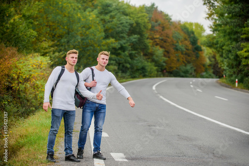 men backpacks walking together by road. twins walking along road. Adventure and discovery. Man with backpack hitchhiking on road. Summer vacation. Tourist traveler travel auto stop. Budget travelers