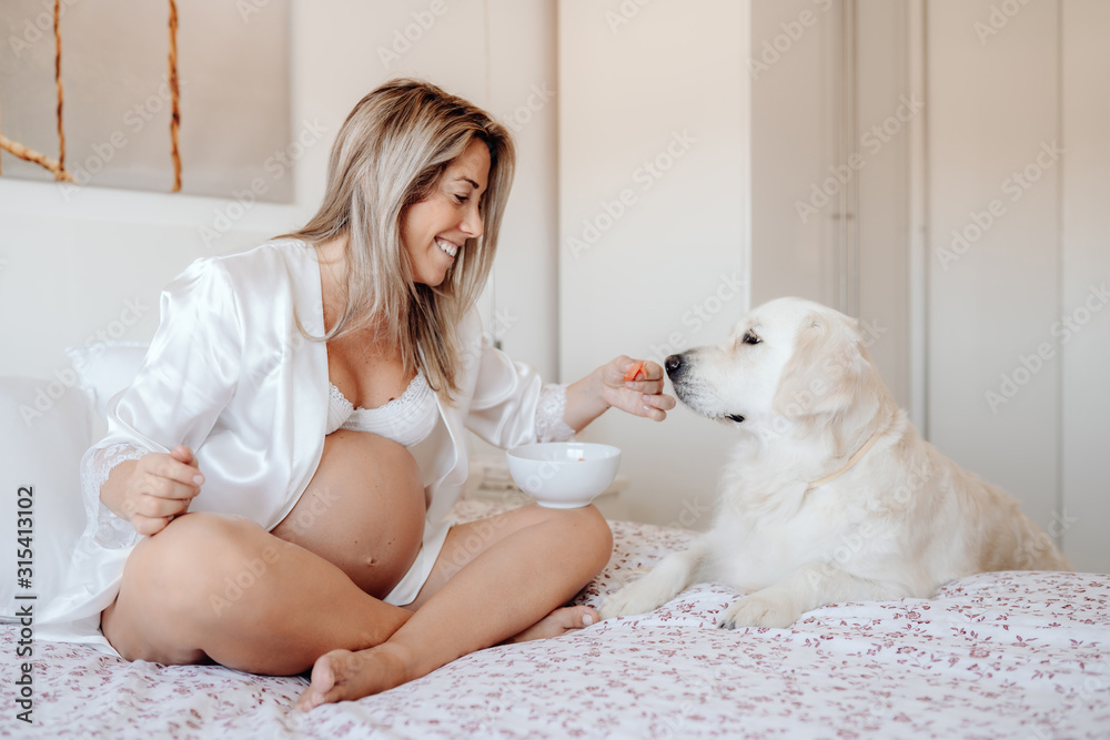 Content blonde pregnant female sitting on bed with crossed legs holding bowl of food while Labrador dog putting paws near and looking at meal Content blonde pregnant female sitting on bed with crossed legs holding bowl of food while Labrador dog putting paws near and looking at meal