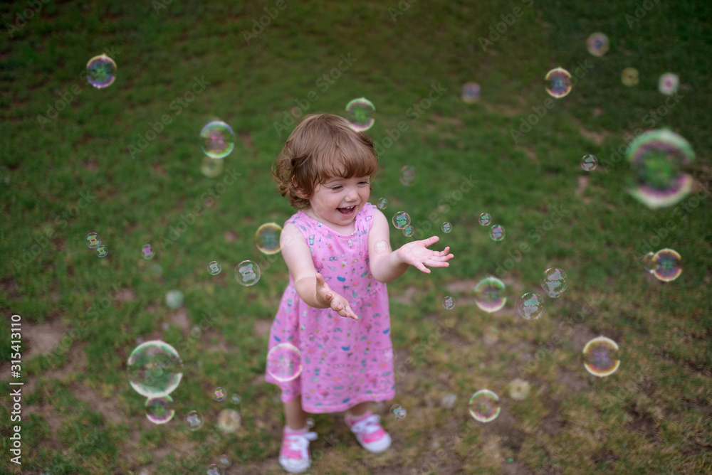 From above adorable child in pink dress laughing and capturing rainbow soap bubbles on green meadow in park From above adorable child in pink dress laughing and capturing rainbow soap bubbles on green meadow in park
